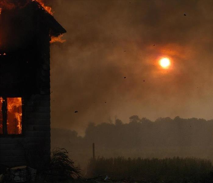 Burning house in left foreground against smoky sky during sunset.